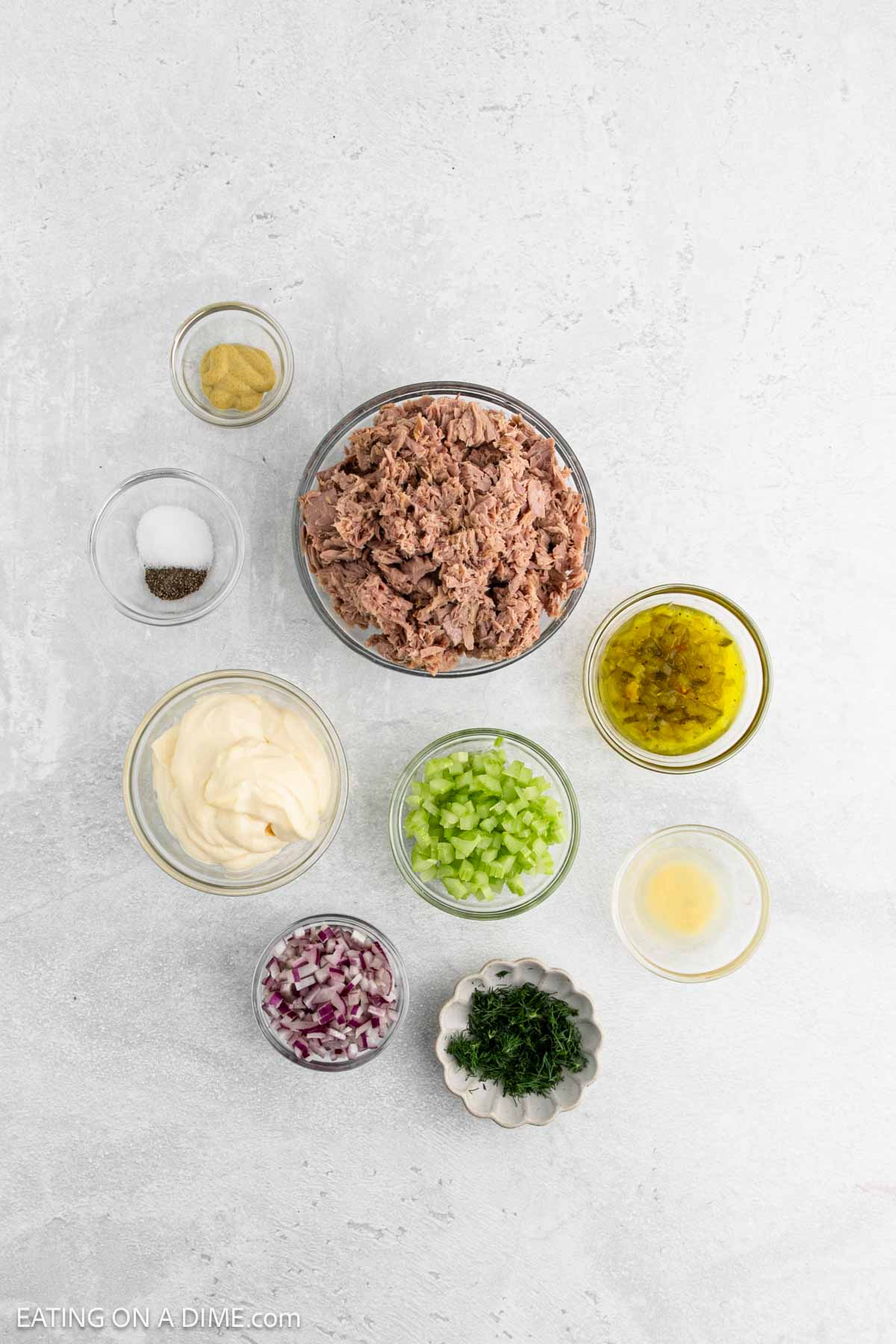 Overhead view of tuna salad ingredients in separate bowls: tuna, mustard, salt and pepper, mayonnaise, chopped celery, chopped red onion, chopped dill, relish, and lemon juice on a light surface.