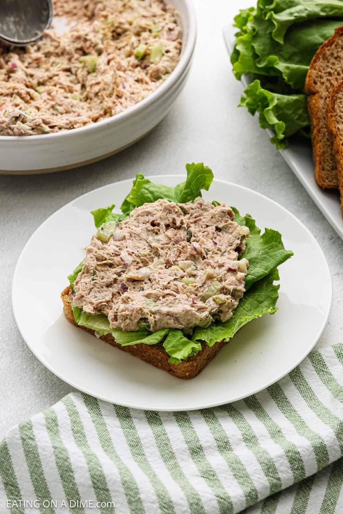 Open-faced tuna salad sandwich with lettuce on whole wheat bread sits on a white plate. A bowl of tuna salad, lettuce leaves, and slices of bread are nearby. A green and white striped towel is in the foreground.