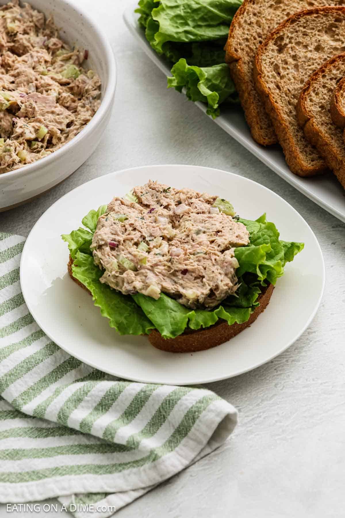 A slice of whole wheat bread topped with leafy lettuce and a scoop of tuna salad on a white plate, next to a striped green towel, with more bread, lettuce, and tuna salad in the background.