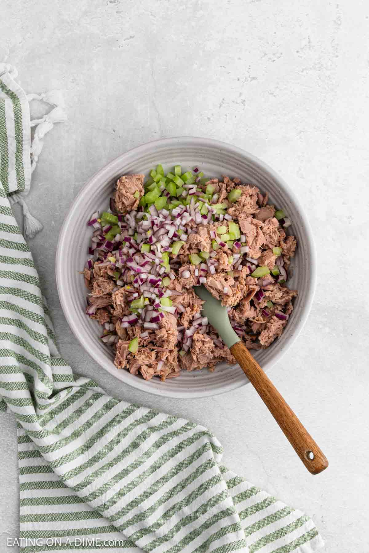 A bowl filled with tuna, chopped red onion, and diced celery, with a wooden spoon inside. A green and white striped kitchen towel is placed next to the bowl on a light-colored surface.