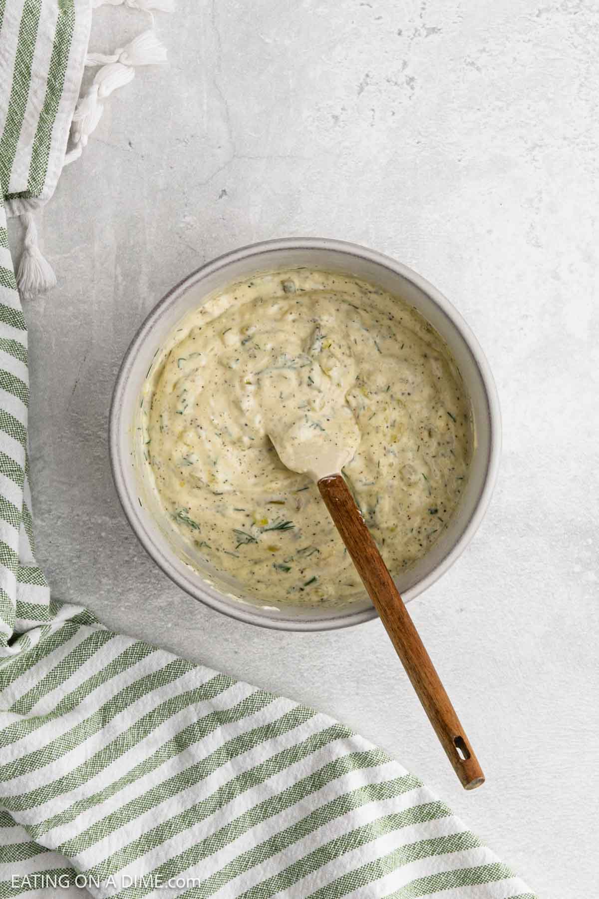 A bowl of creamy homemade tzatziki sauce with visible herbs, stirred by a wooden-handled spoon, sits on a light gray surface next to a green and white striped towel.