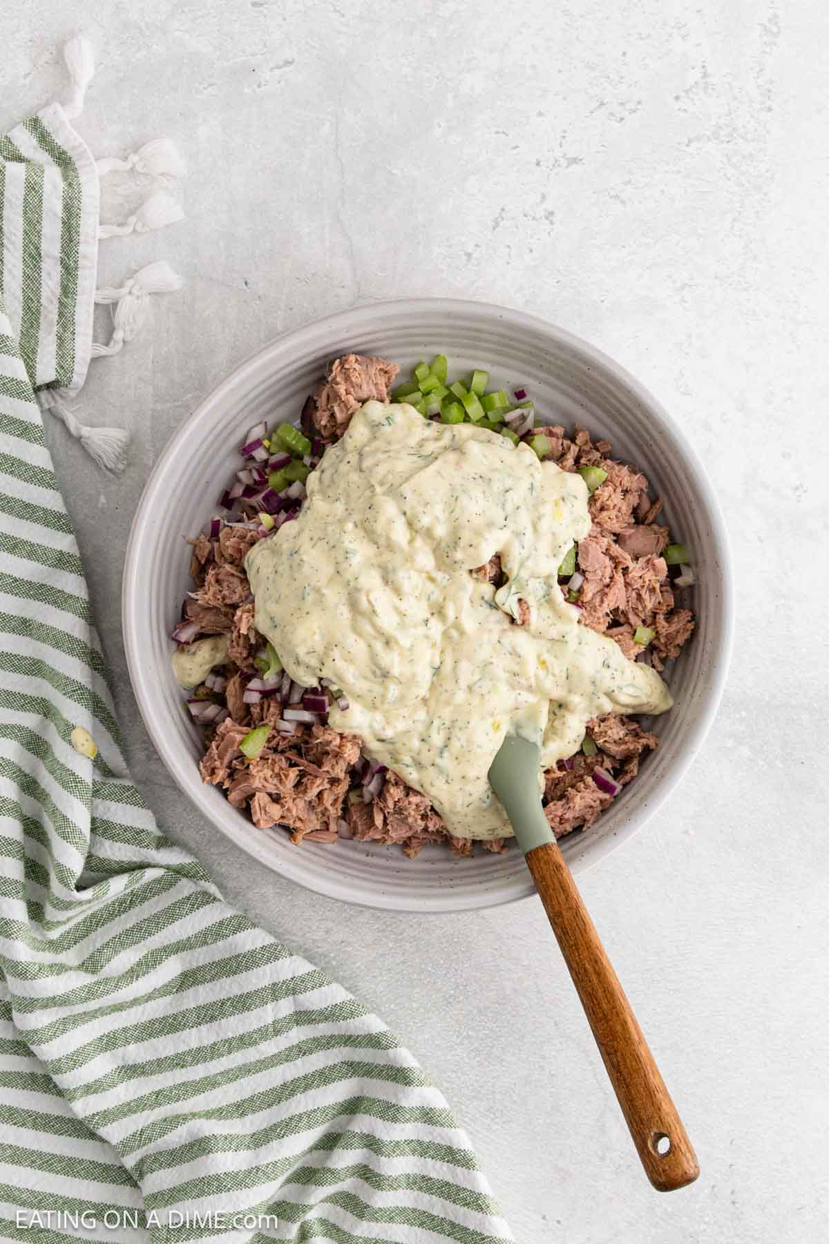 A bowl with chopped celery, red onion, canned tuna, and a creamy dressing, all ready to be mixed. A spatula rests in the bowl and a green-and-white striped towel is nearby.