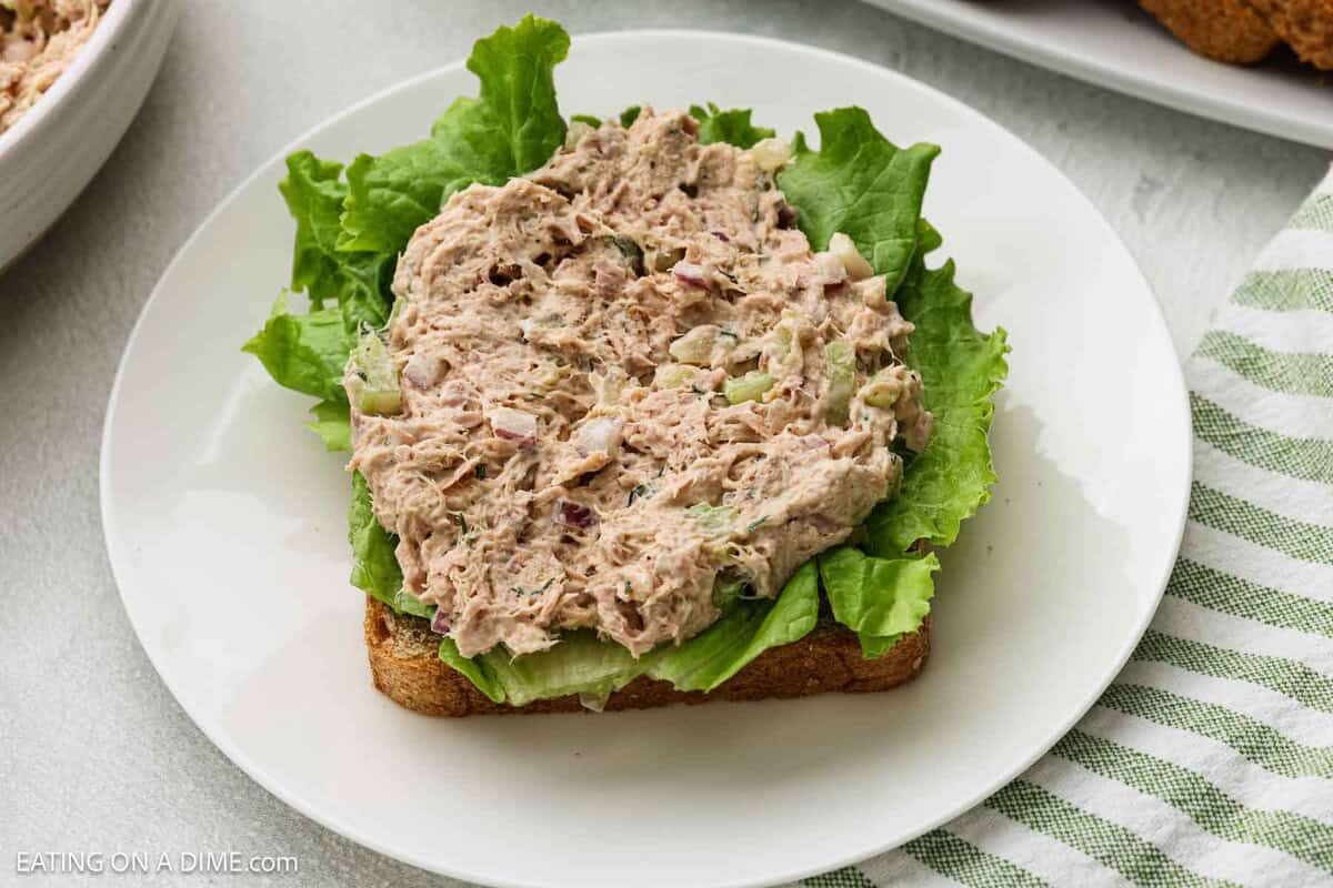 An open-faced sandwich with leafy lettuce and a generous scoop of tuna salad on a slice of whole grain bread, served on a white plate with a striped green and white napkin nearby.