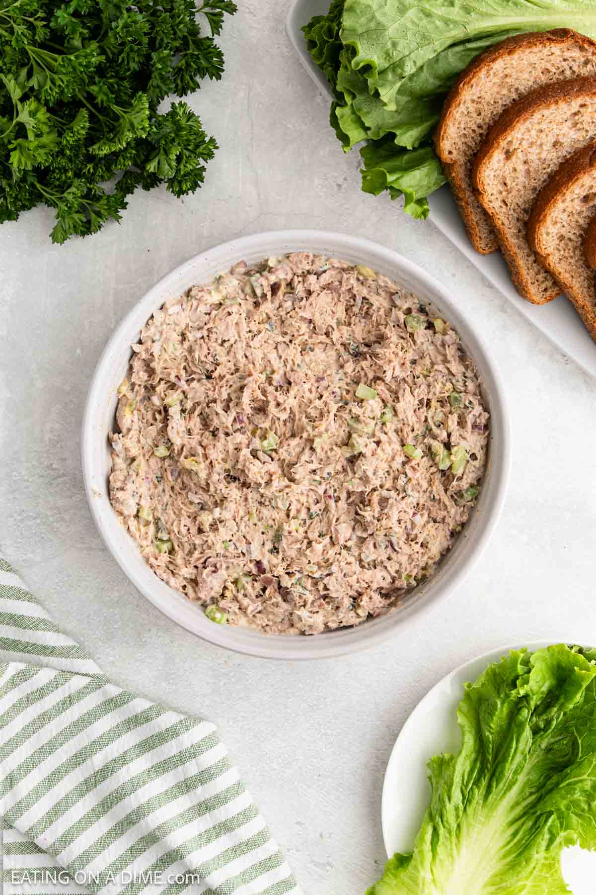 A bowl of tuna salad on a light surface, surrounded by fresh parsley, lettuce leaves, slices of whole wheat bread, and a green-striped kitchen towel.