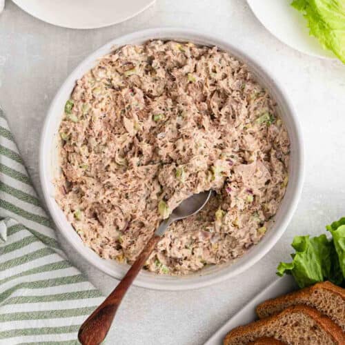 A bowl of tuna salad mixed with chopped celery and red onion, with a spoon in the bowl. Slices of bread, a striped towel, and lettuce leaves are nearby on a light-colored surface.