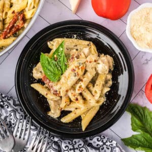 A black plate filled with creamy Chicken Pesto Pasta, sun-dried tomatoes, and a fresh basil leaf, surrounded by grated cheese, tomatoes, and utensils on a patterned napkin.