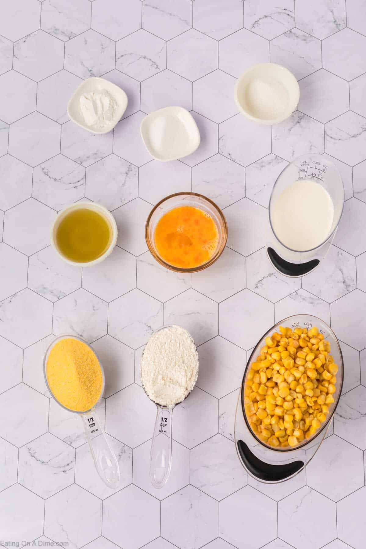 A top view of various ingredients for a Corn Fritters recipe in small bowls and measuring cups on a white hexagon-tiled surface, including corn, flour, cornmeal, oil, egg, milk, and seasonings—perfect for delicious corn recipes.