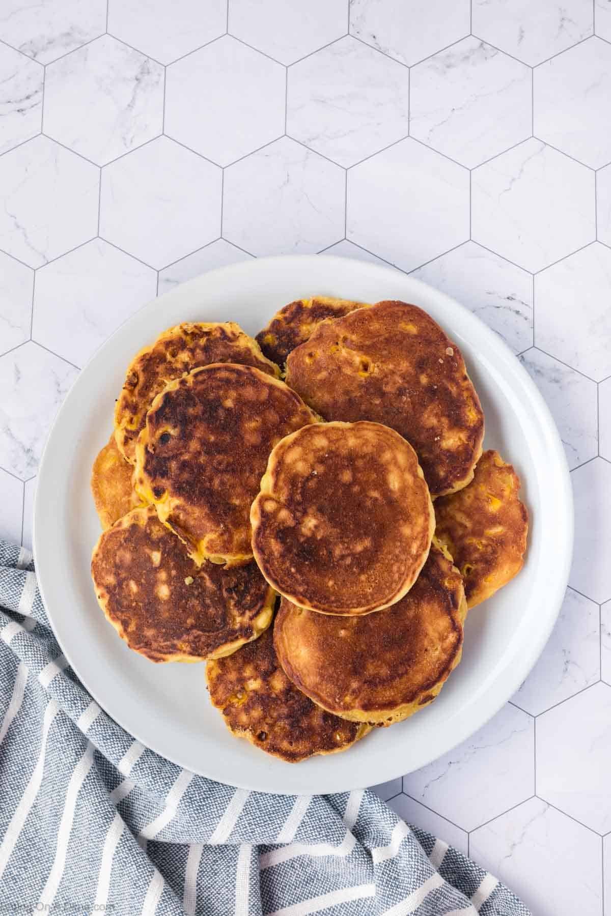 A plate of golden-brown corn fritters is placed on a white hexagonal-tile surface, with a blue and white striped cloth partially visible beside the plate.