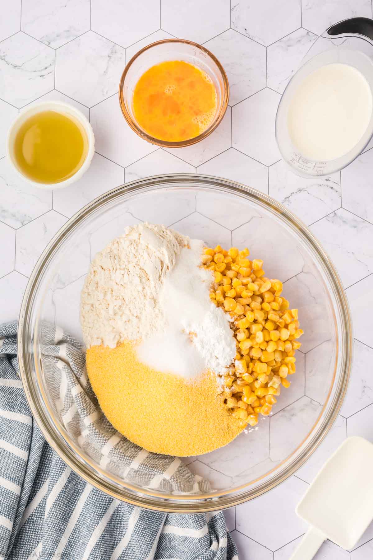 A large glass bowl holds cornmeal, flour, sugar, salt, and corn kernels—perfect for making corn fritters. Nearby are bowls of melted butter, beaten eggs, and a pitcher of milk on a white hexagonal tile surface with a striped towel.