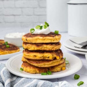 A stack of four golden corn fritters, a classic among Corn Recipes and Easy Appetizers, is topped with sour cream and chopped green onions on a white plate, with a blue-striped napkin and extra fritters in the background.