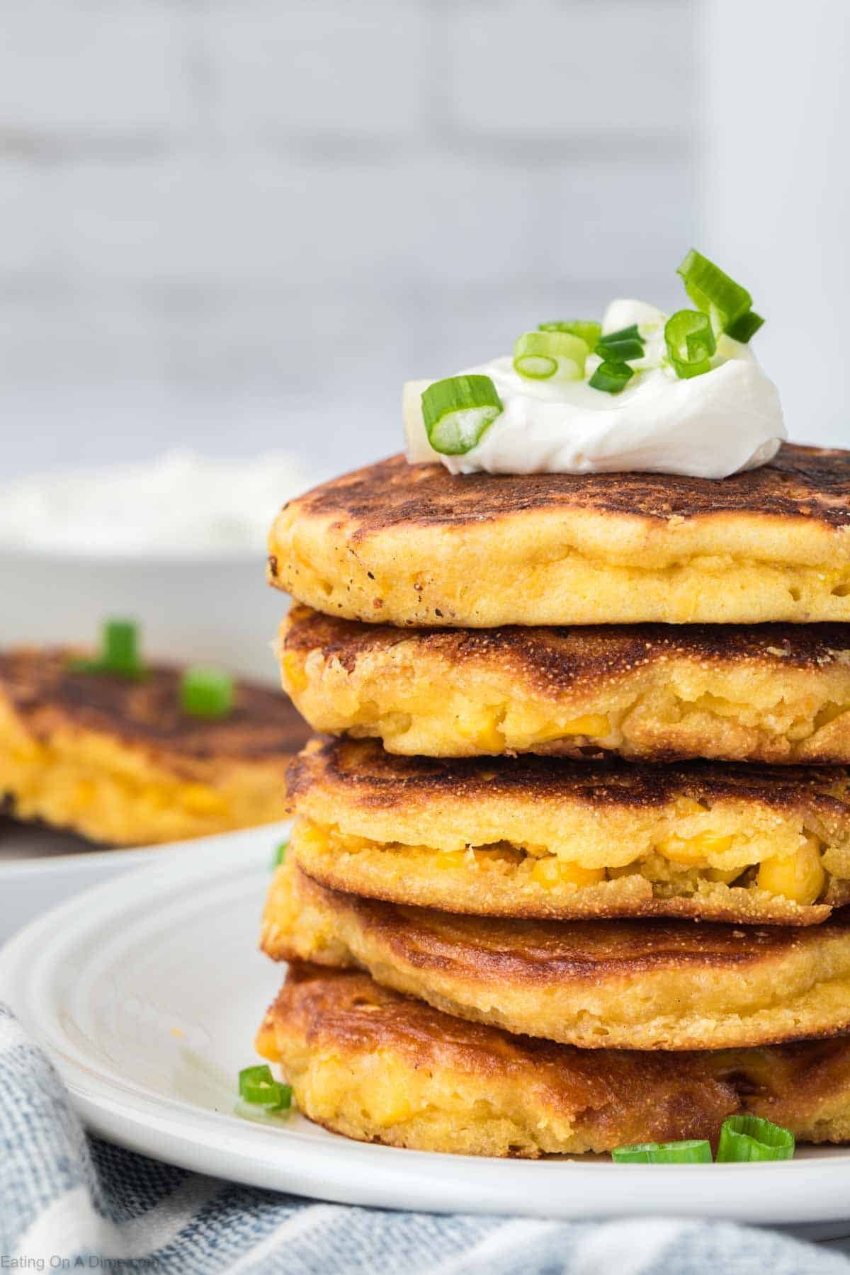 A stack of golden, crispy corn fritters—one of the best easy appetizers—topped with sour cream and chopped green onions sits on a white plate, with more fritters blurred in the background.