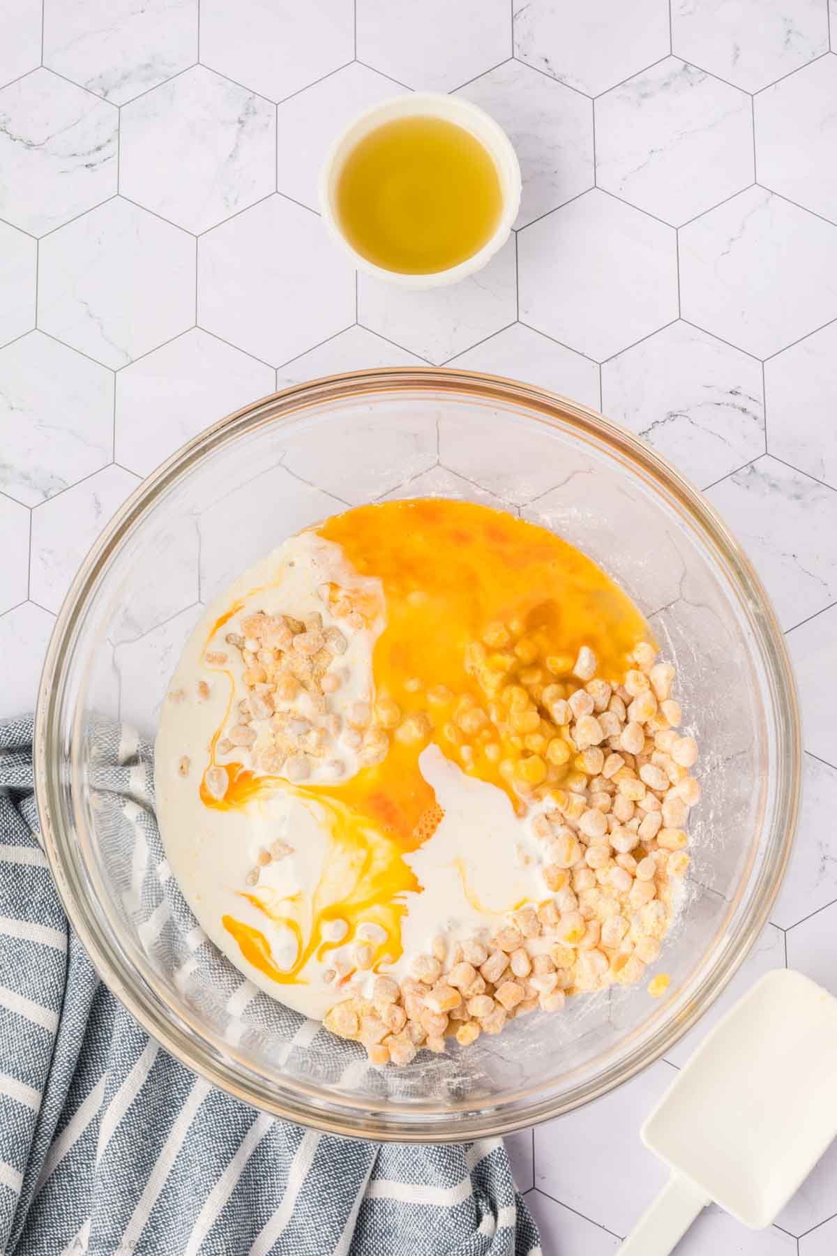 A glass bowl with flour, egg yolks, chopped nuts, and milk sits on a white hexagon-tiled surface—ingredients ready to make delicious corn fritters—next to a small bowl of oil, a spatula, and a gray-striped cloth.