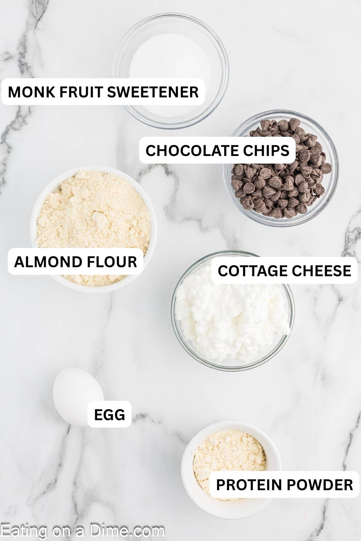 Five glass bowls hold sugar, chocolate chips, almond flour, cottage cheese, and more almond flour on a marble surface.