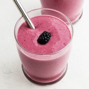 A glass filled with a thick, pink cottage cheese smoothie, topped with a single blackberry and a metal straw, sits on a light surface. Another similar glass is partially visible in the background.