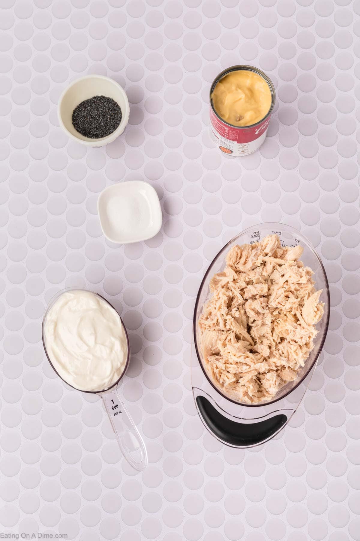 Overhead view of shredded chicken in a bowl, a measuring cup of sour cream, poppy seeds, salt, and an open can of cream soup—perfect ingredients for a classic Poppyseed Chicken Casserole on a patterned surface.