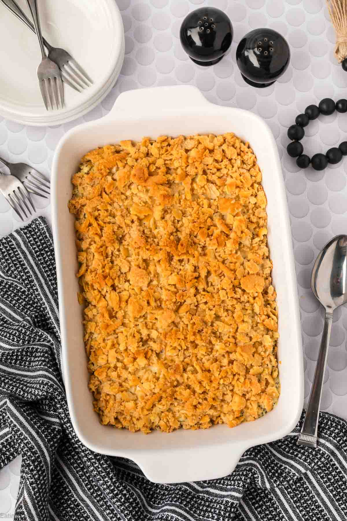 A casserole dish filled with a golden, crunchy topping sits on a white surface—this classic Poppyseed Chicken casserole is ready to serve. Forks, plates, a striped towel, and black salt and pepper shakers are arranged nearby.