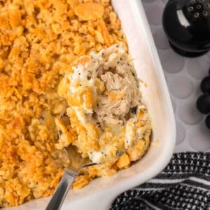 A close-up of a serving spoon lifting creamy Poppyseed Chicken casserole with a golden, crunchy cracker topping from a white baking dish. A black pepper shaker and a striped cloth are nearby.