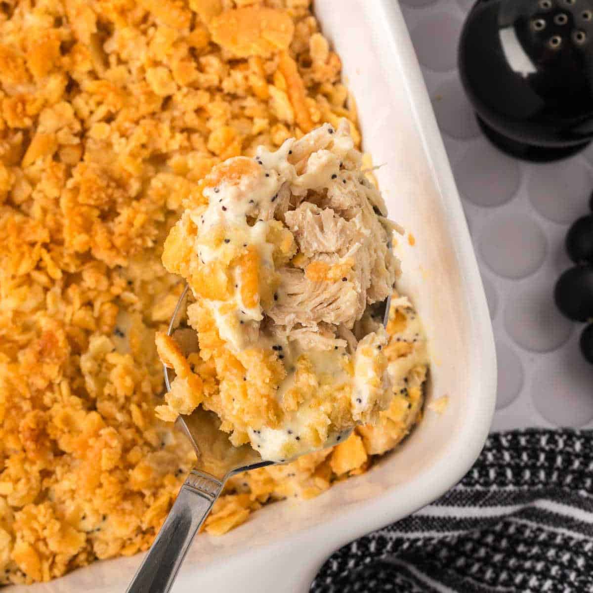 A close-up of a serving spoon lifting creamy Poppyseed Chicken casserole with a golden, crunchy cracker topping from a white baking dish. A black pepper shaker and a striped cloth are nearby.