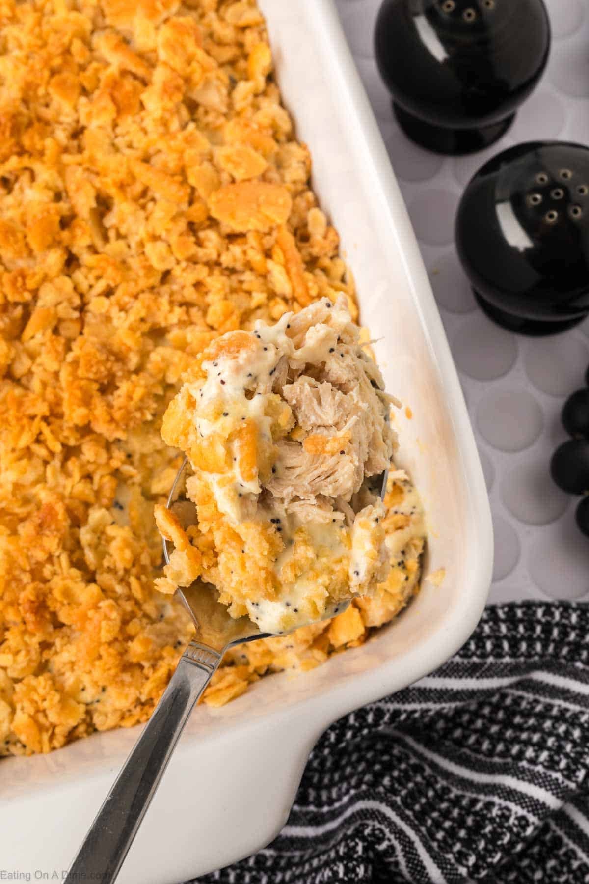 A close-up of a casserole dish with a golden, crumbly topping, a serving spoon lifting out creamy Poppyseed Chicken filling. Black salt and pepper shakers and a striped cloth are nearby—perfect for fans of poppy seed recipes.