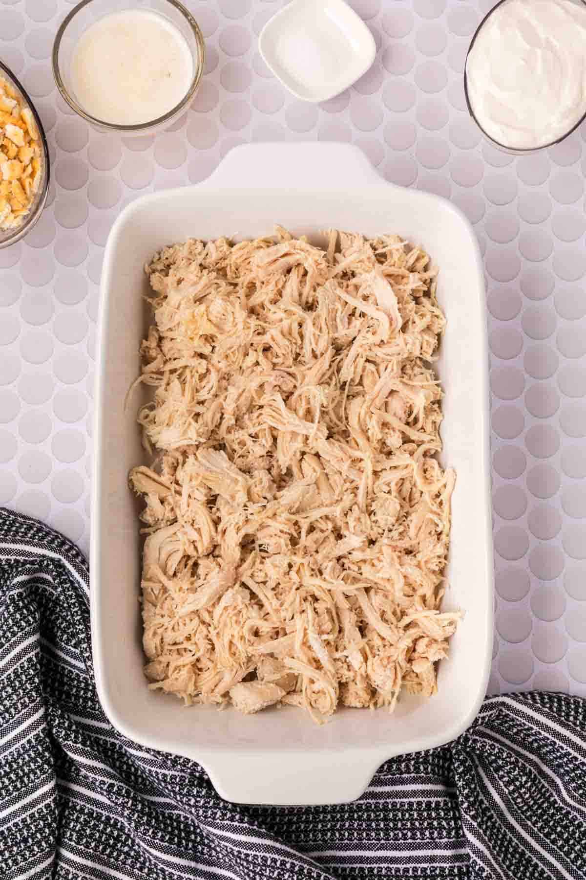 A white baking dish filled with shredded chicken for an Easy Chicken Recipe sits on a light textured surface. Surrounding the dish are small bowls of sauce, cheese, and seasoning, with a black and white striped towel in the lower left corner.