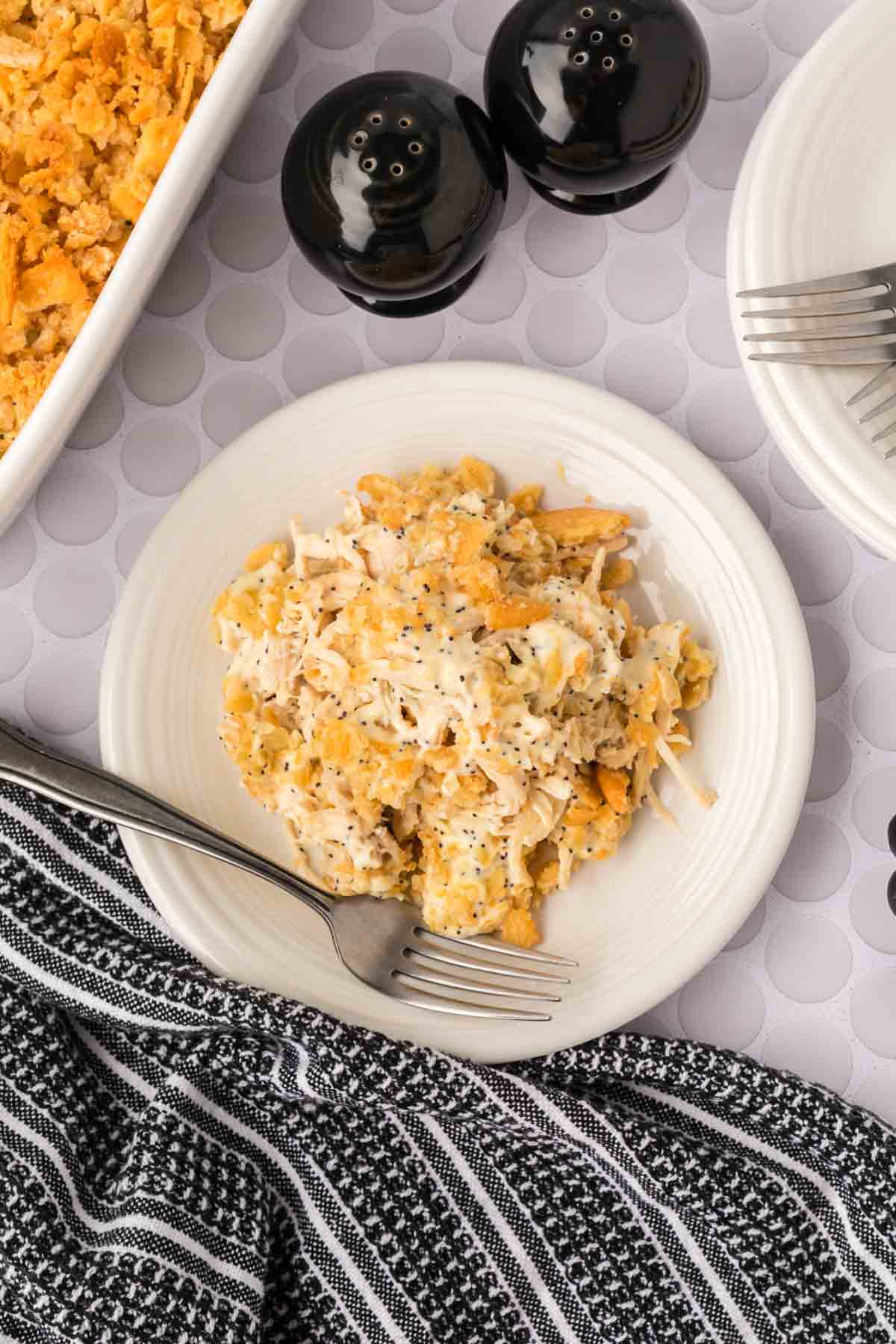 A white plate with a serving of cheesy Poppyseed chicken casserole, topped with poppy seeds, and a fork resting on the plate. Nearby are a black-and-white striped napkin, salt and pepper shakers, and part of the casserole dish.