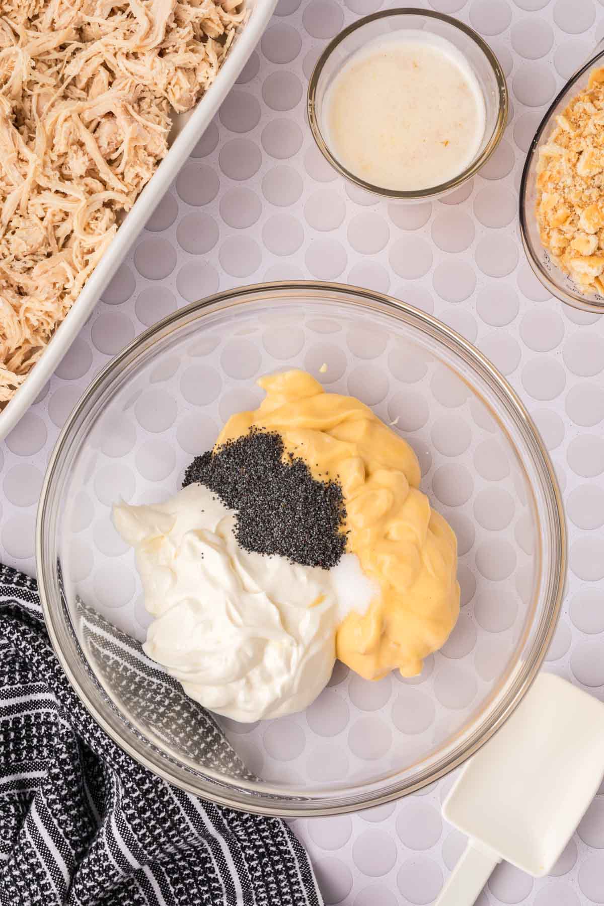 A glass bowl with mayonnaise, sour cream, yellow sauce, and poppy seeds sits on a counter next to shredded chicken—perfect for making Poppyseed Chicken or your favorite poppy seed recipe—alongside melted butter, crushed crackers, and a spatula.