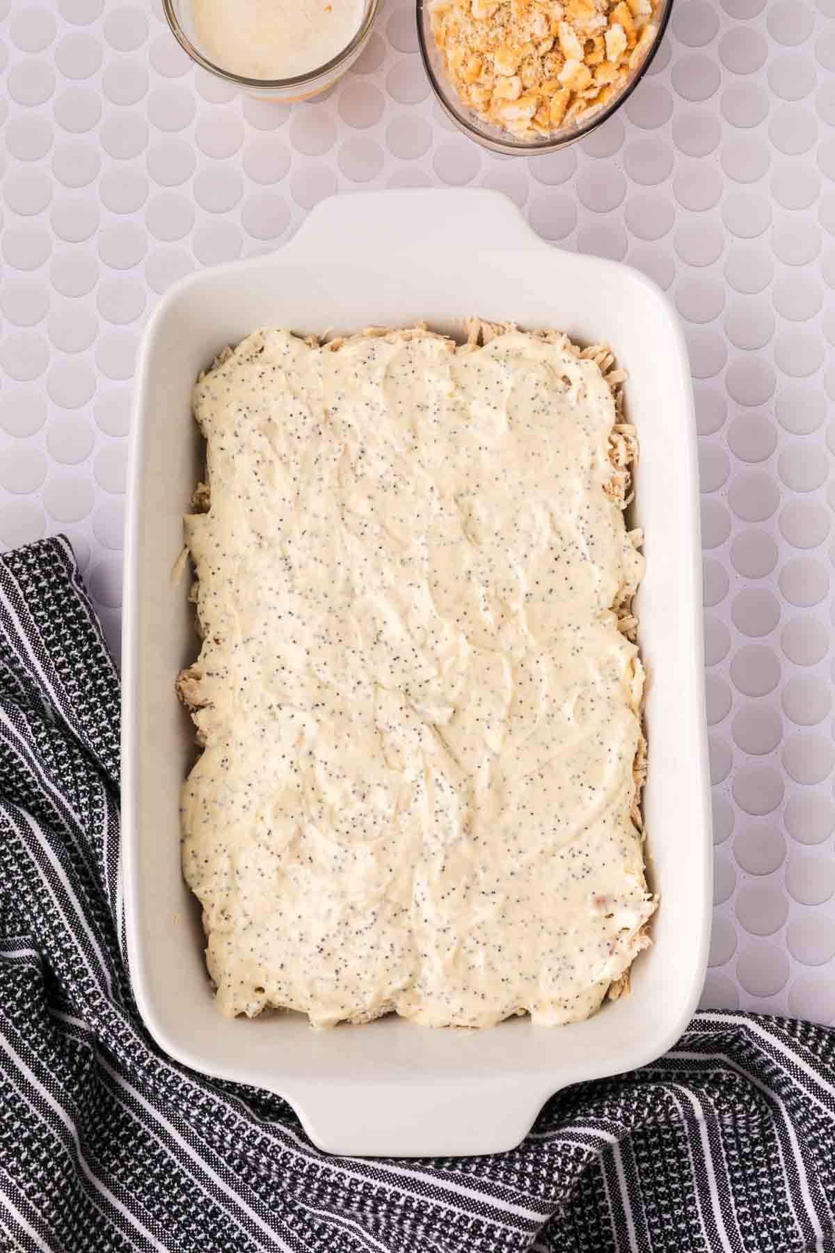 A white baking dish filled with creamy Poppyseed Chicken casserole sits on a white textured surface with a black and white striped towel beside it. Two small bowls are at the top of the image.