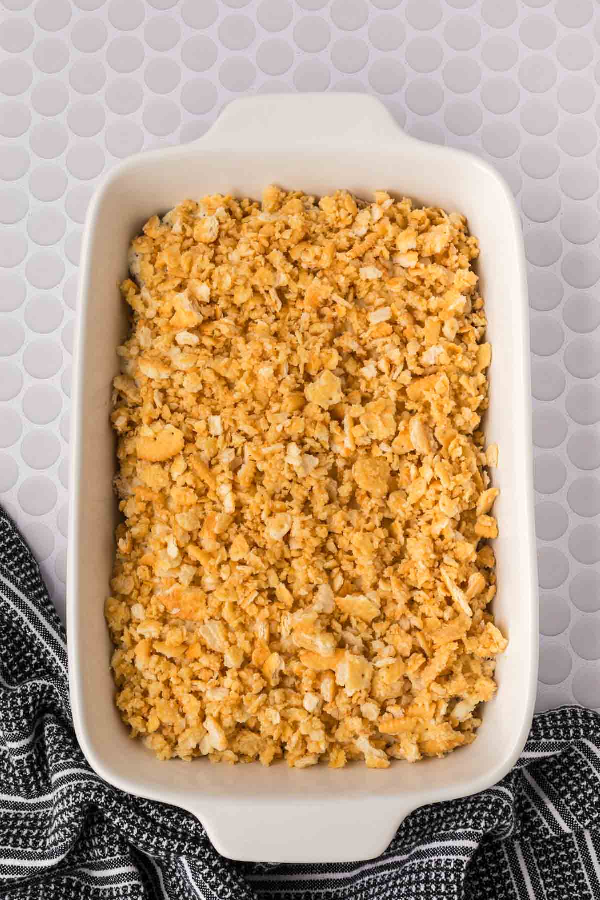 A rectangular white baking dish filled with a golden, cracker crumb topping mixture sits on a light patterned surface next to a black and white striped cloth—perfect for serving classic Poppyseed Chicken casserole.