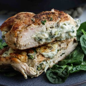 Spinach Stuffed Chicken Breast: Two golden brown chicken breasts filled with creamy cheese and fresh spinach, stacked on a dark plate with extra spinach leaves, perfectly seasoned with herbs.