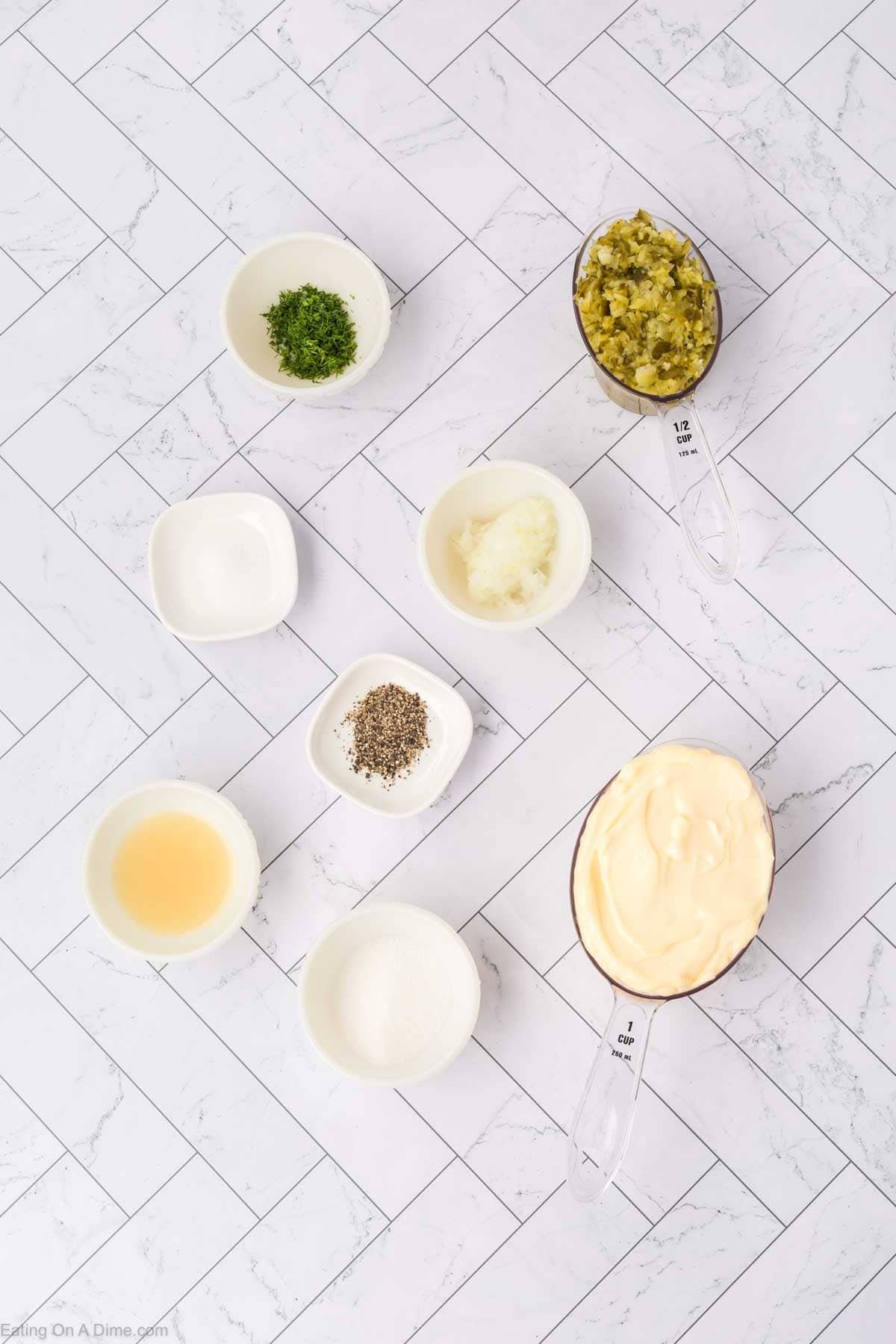 Eight small bowls and measuring cups holding ingredients for homemade tartar sauce—mayonnaise, chopped herbs, relish, diced onions, pepper, and liquids—are arranged on a white tiled surface.