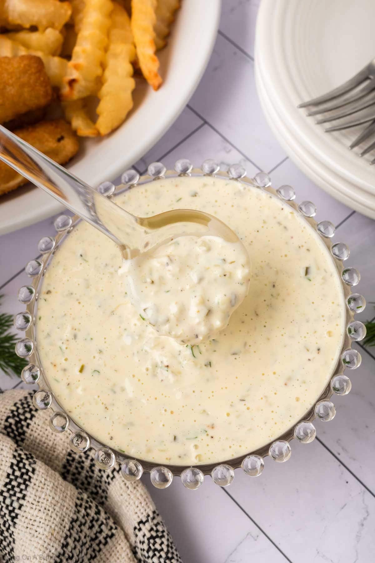 A glass bowl filled with creamy Tartar Sauce, made from a classic Tartar Sauce Recipe and garnished with herbs, sits on a table next to crinkle-cut fries and breaded fish. A spoon rests in the sauce, ready for serving.