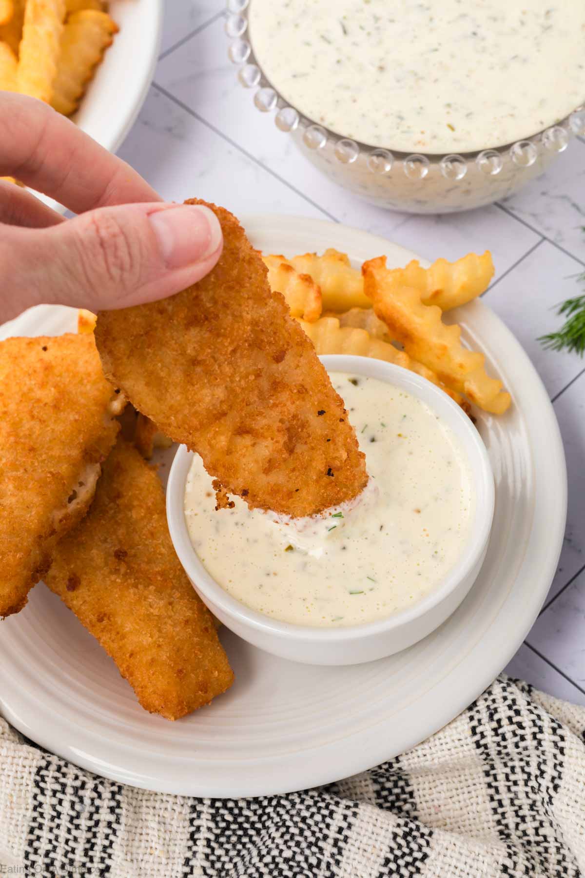 A hand dips a crispy breaded fish fillet into a bowl of homemade tartar sauce on a plate with crinkle-cut fries; more sauce and fries are visible in the background.