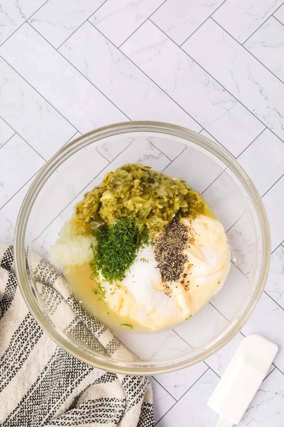 A glass bowl on a tiled surface contains Homemade Tartar Sauce with mayonnaise, chopped pickles, onions, dill, pepper, and other seasonings. A striped kitchen towel and a white measuring spoon sit nearby.