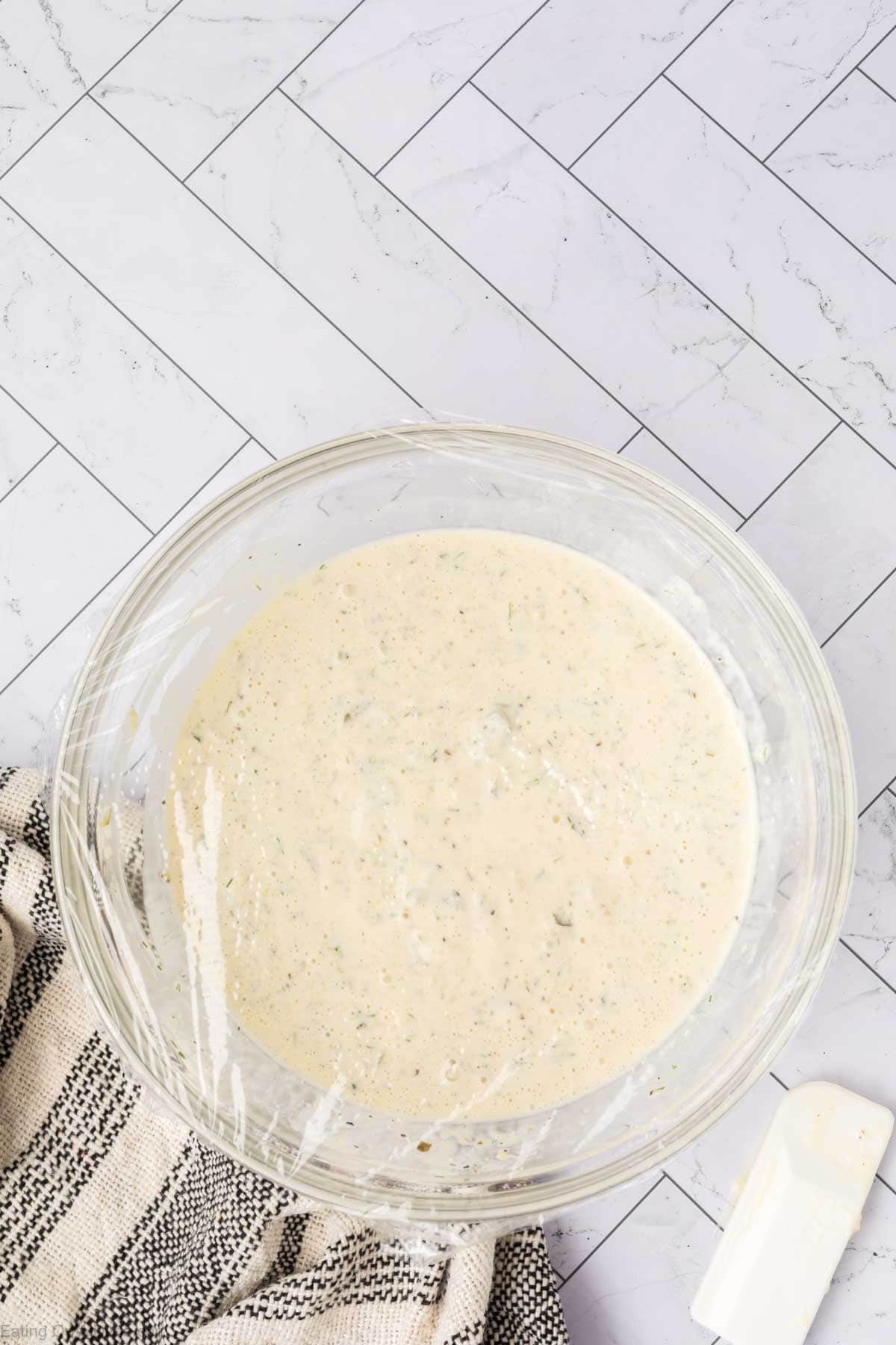 A glass bowl filled with creamy, homemade tartar sauce speckled with herbs sits on a striped kitchen towel atop a white tiled surface; a white spatula is nearby. Try this classic tartar sauce recipe for your next meal.