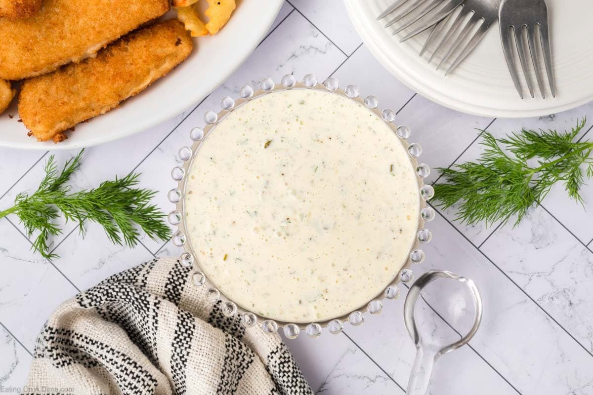 A bowl of creamy Homemade Tartar Sauce sits on a tiled surface, surrounded by fresh dill, a folded kitchen towel, a plate of breaded fish fillets, and a stack of white plates with forks.