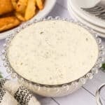 A glass bowl of creamy tartar sauce, speckled with herbs, sits on a white surface near fried food, a striped cloth, and stacked white plates with forks—perfect for trying your favorite tartar sauce recipe.