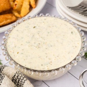 A glass bowl of creamy tartar sauce, speckled with herbs, sits on a white surface near fried food, a striped cloth, and stacked white plates with forks—perfect for trying your favorite tartar sauce recipe.