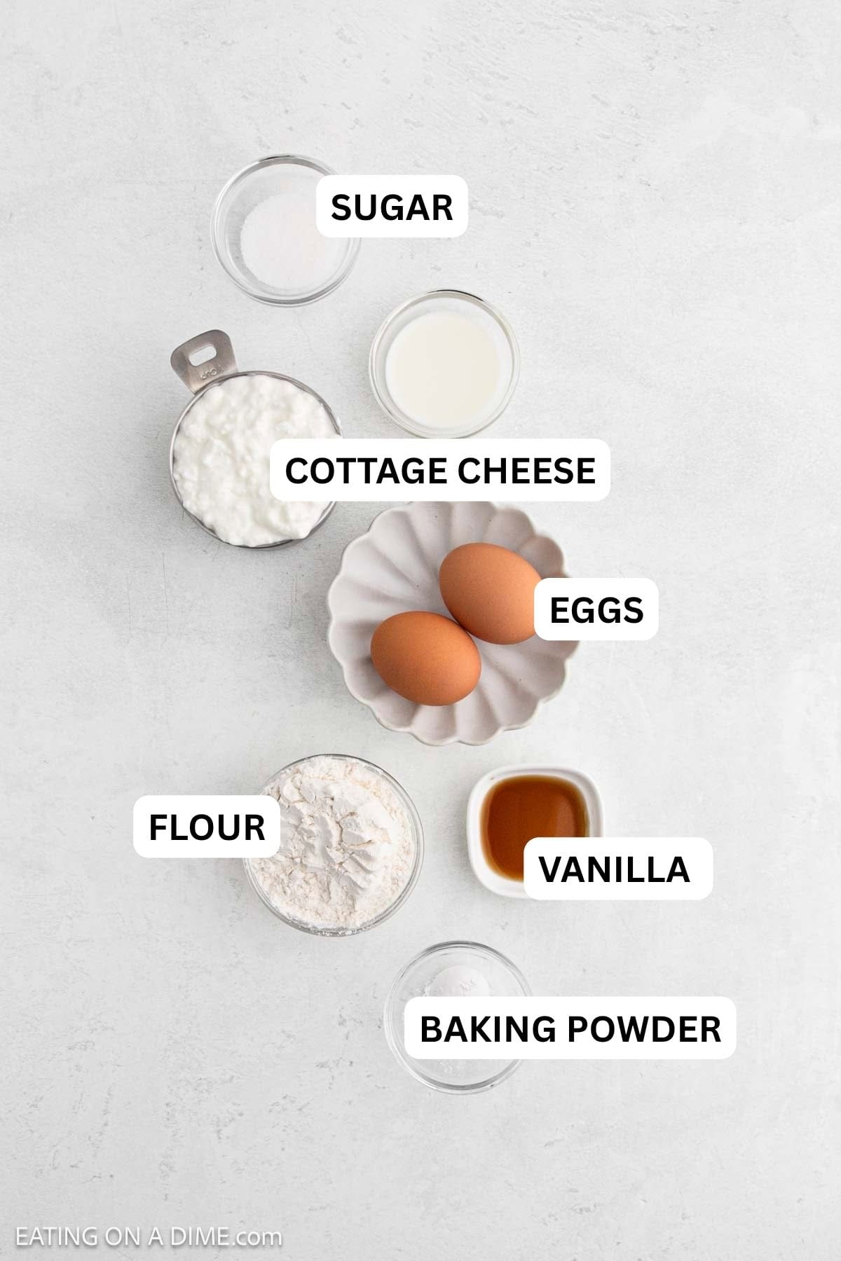 Ingredients needed for this recipe includes two eggs in a small dish, cottage cheese, flour, milk, sugar, vanilla extract, and baking powder.
