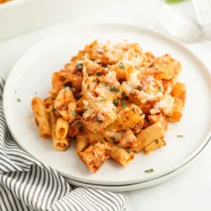 A serving of baked ziti with marinara sauce, pieces of chicken, melted cheese, and fresh herbs on a white plate, part of our Meal Plan 55, sits next to a striped cloth napkin.