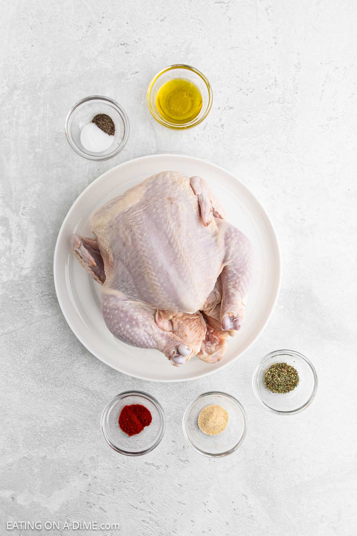 A raw Air Fryer Whole Chicken rests on a white plate, surrounded by small bowls of olive oil, salt and pepper, paprika, garlic powder, and dried herbs on a light gray surface.