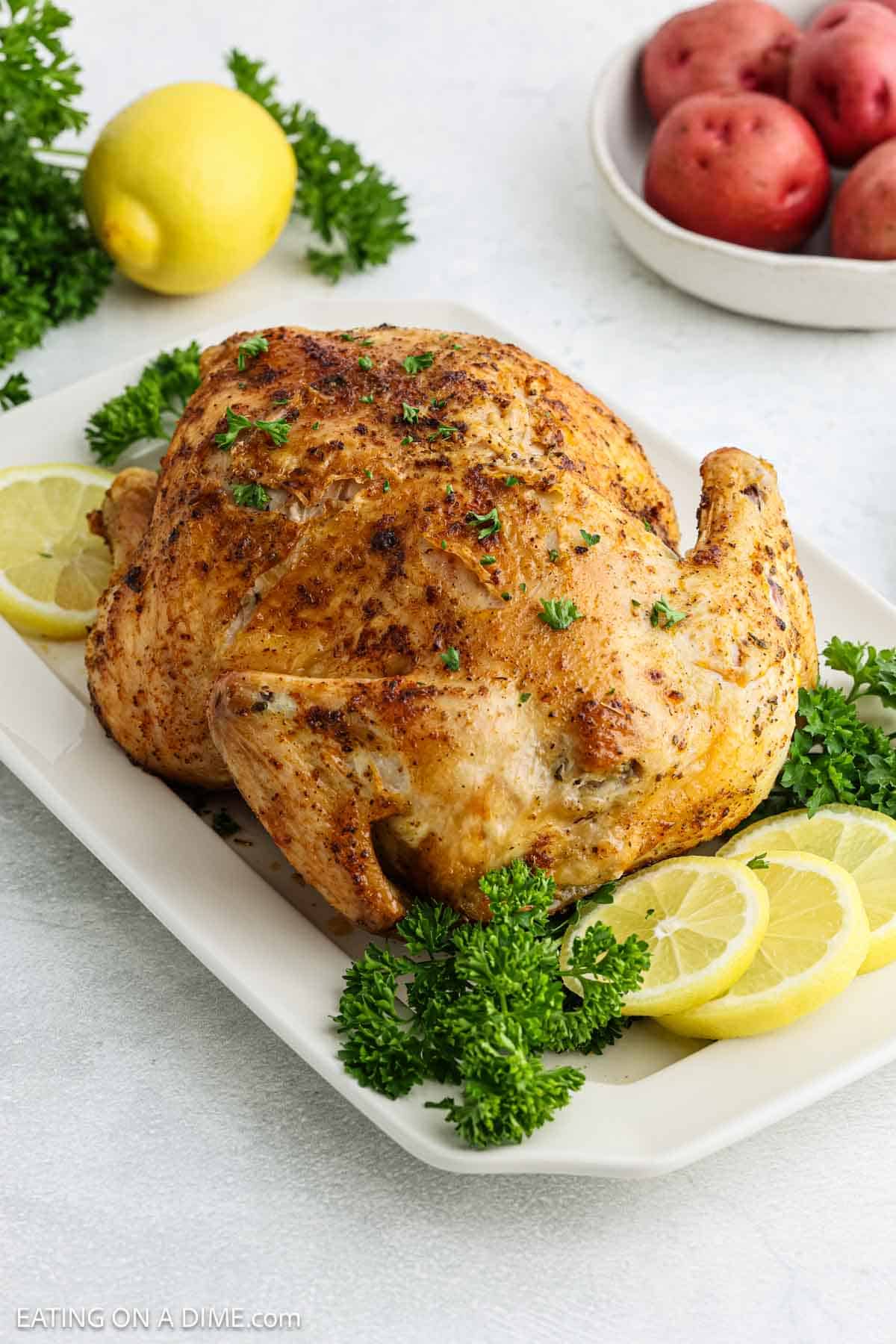 A whole roasted Air Fryer Whole Chicken garnished with parsley and lemon slices on a white platter. In the background, there is a lemon, a bowl of red potatoes, and more parsley.
