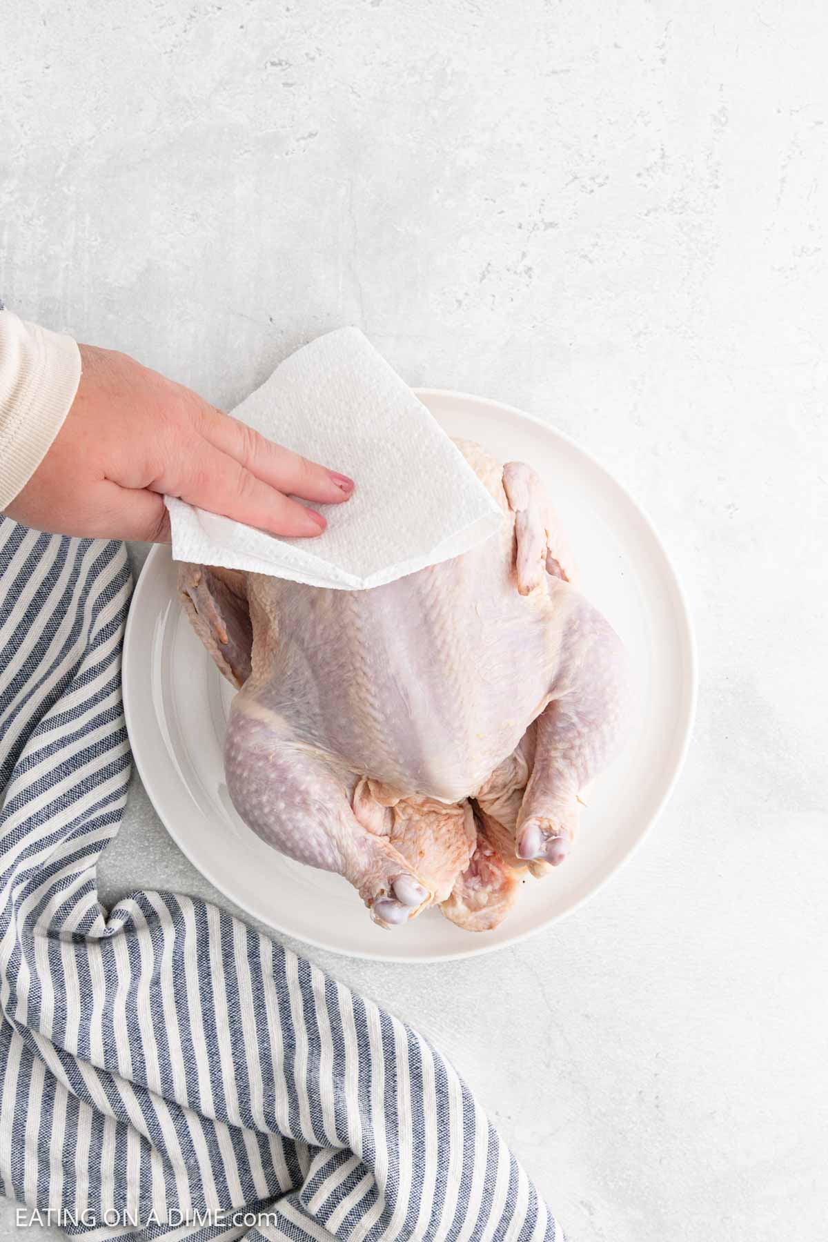 A hand uses a paper towel to pat dry a raw whole chicken on a white plate, prepping it for Air Fryer Whole Chicken, with a striped kitchen towel nearby on a light surface.