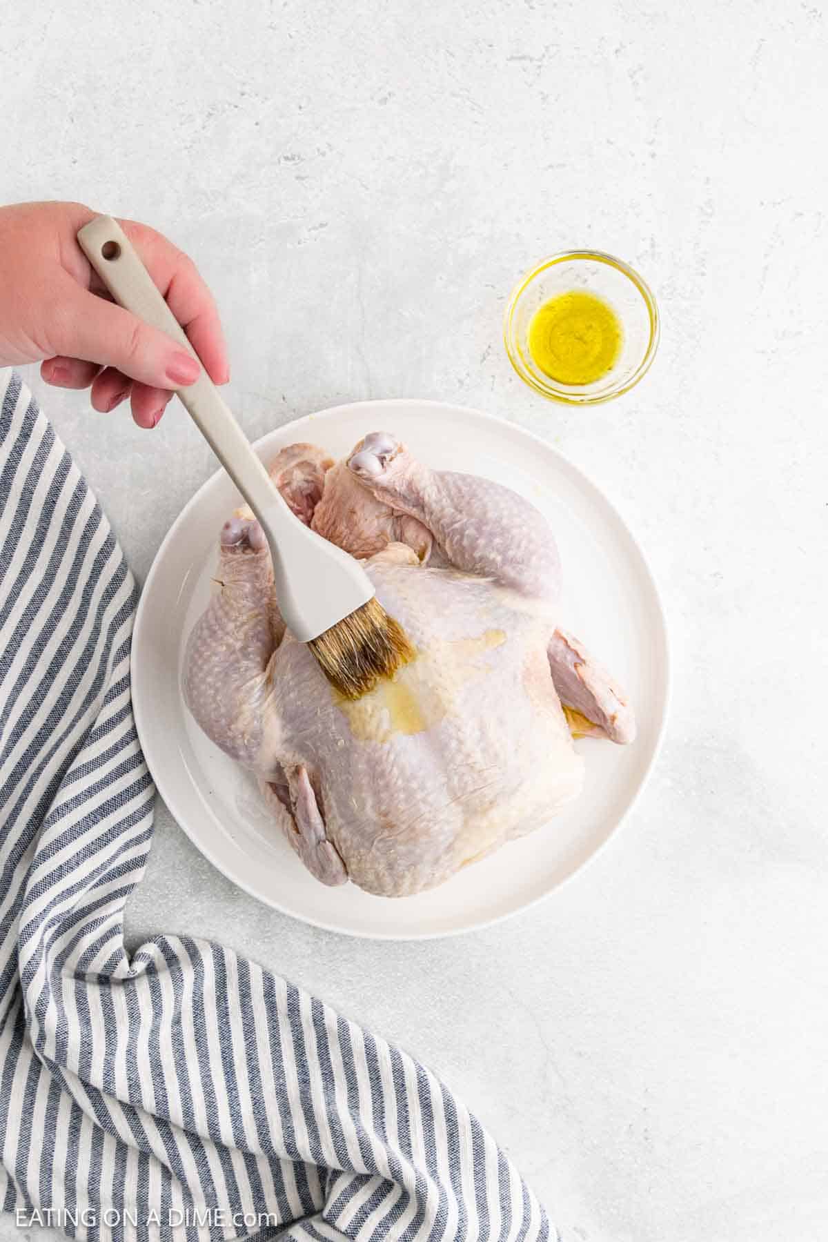 A hand uses a brush to apply oil to a raw whole chicken on a white plate, prepping it as an air fryer whole chicken. A small bowl of oil sits nearby, with a blue-striped cloth to the side.