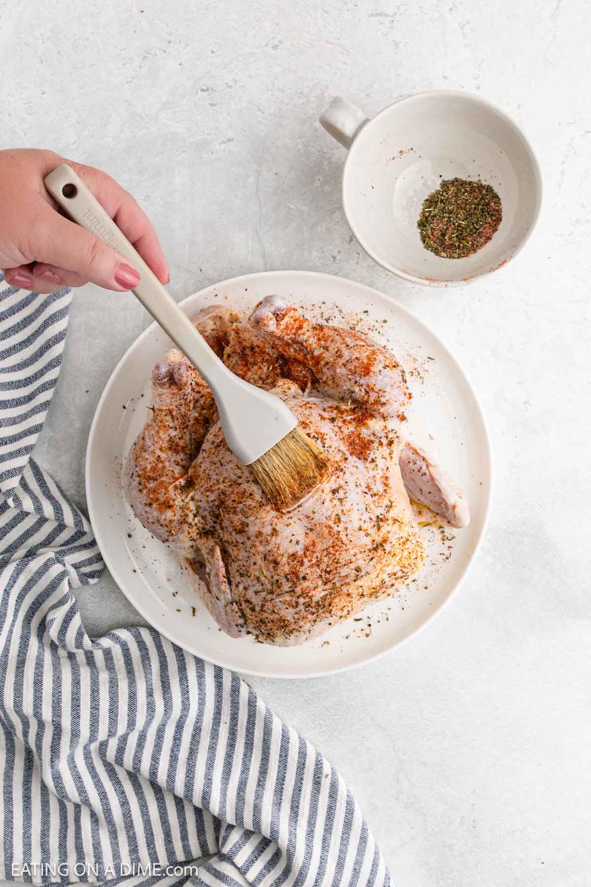 A hand uses a brush to apply seasoning to a raw whole chicken on a white plate. A bowl of mixed spices and a striped kitchen towel sit nearby, ready for preparing this Air Fryer Whole Chicken on a light-colored surface.