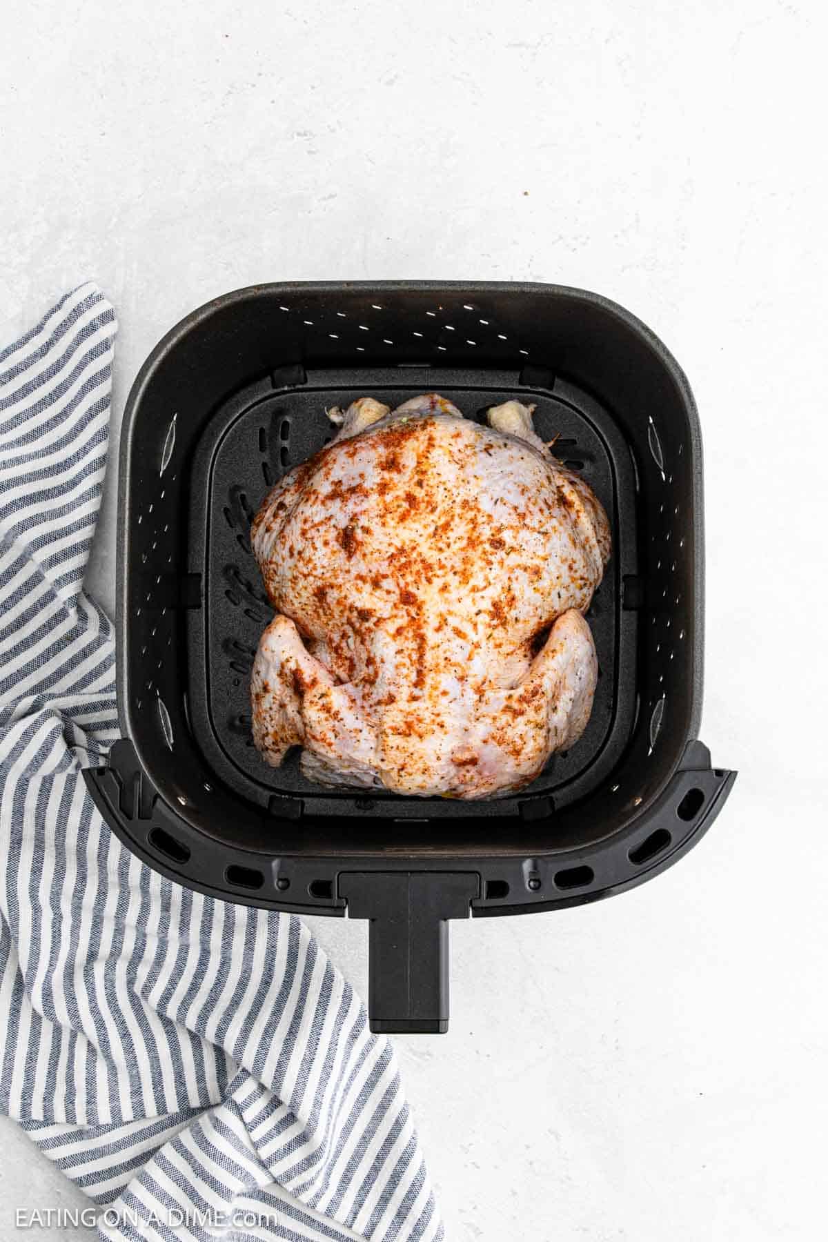 A seasoned Air Fryer Whole Chicken sits inside an air fryer basket on a white surface, next to a blue and white striped kitchen towel.