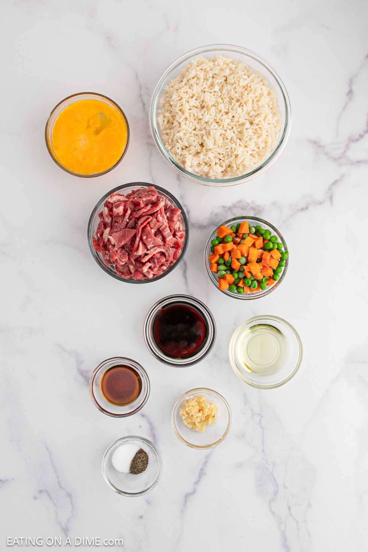 Nine bowls on a white marble surface showcase the ingredients for Beef Fried Rice: beaten eggs, cooked rice, sliced beef, mixed peas and carrots, soy sauce, oil, garlic, plus small dishes of salt, pepper, and brown sauce.