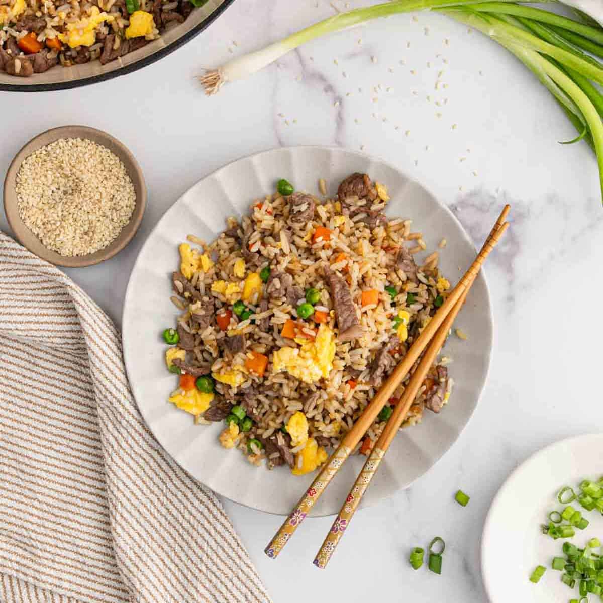 A plate of Beef Fried Rice with eggs, peas, and carrots is served with chopsticks. Sesame seeds, green onions, and a striped napkin rest nearby on a white marble surface.