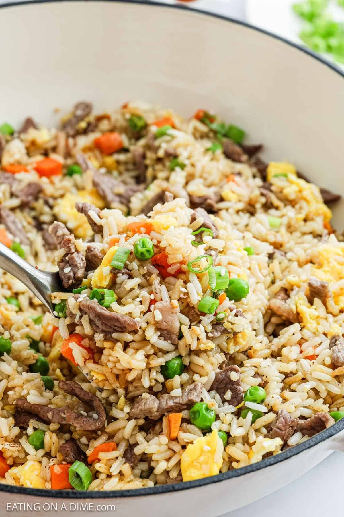 A close-up of a spoonful of Beef Fried Rice with peas, carrots, scrambled eggs, and green onions in a white pot.