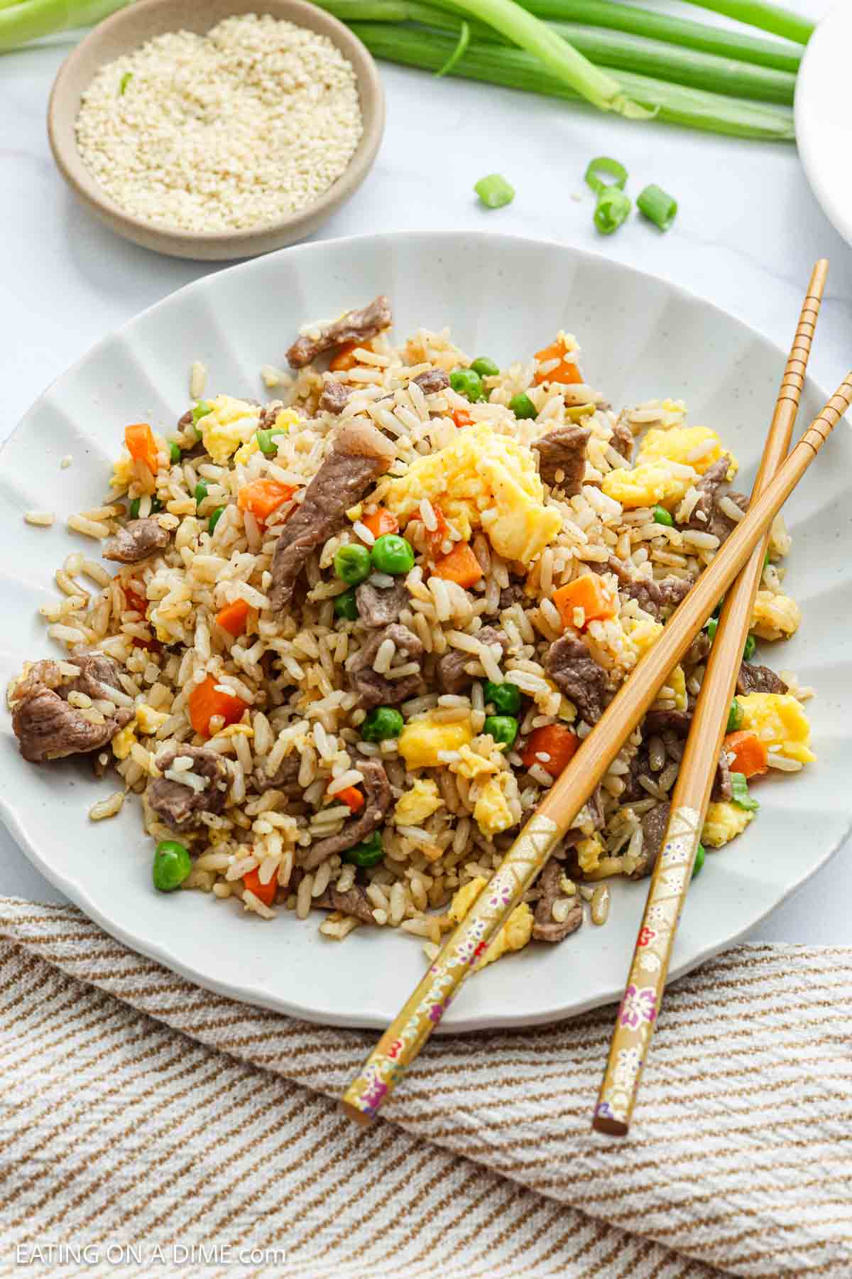 A plate of Beef Fried Rice with peas, carrots, scrambled eggs, and green onions is served with chopsticks. In the background, a bowl of sesame seeds and fresh green onions adds the perfect touch to this savory dish.