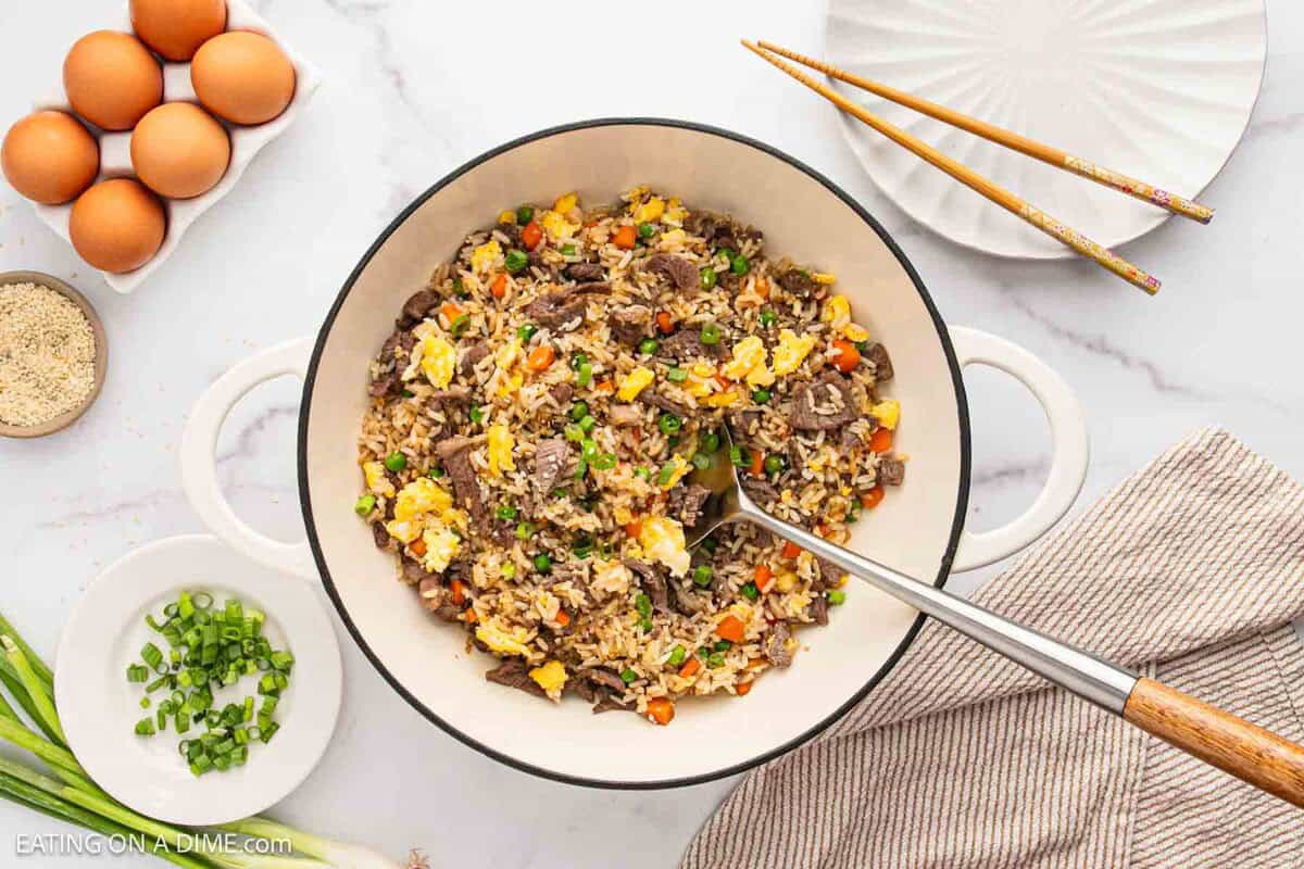 A large white pot filled with delicious Beef Fried Rice with vegetables and scrambled eggs, surrounded by fresh green onions, eggs, sesame seeds, a plate with chopsticks, and a fork on a striped napkin. A flavorful beef recipe to savor.